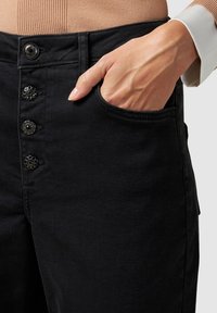 Pantalones de mezclilla negros con cintura alta, cierre de cinco botones con botones decorativos negros y bolsillos laterales.