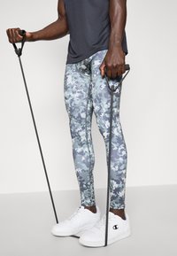 Színes álcázó leggings kék és szürke árnyalatokban, sötét atlétatrikóval és fehér sportcipővel párosítva. Rugalmas ellenállásgumikat tart.
