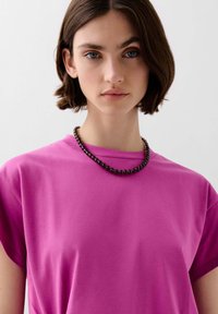 Roze t-shirt met korte mouwen van katoen, met een ronde halslijn. Het model draagt een zwarte kralenketting als accentdetail.