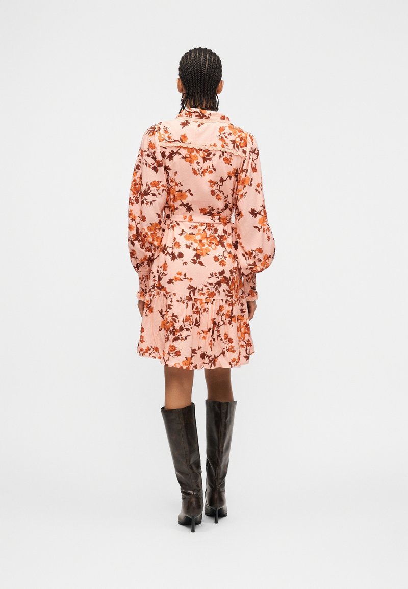 Personne aux cheveux tressés portant une robe rose fleurie à manches longues et des bottes noires montantes à talons hauts, debout de dos sur un fond blanc.