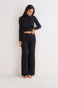 Vrouw die blootsvoets op een tapijt staat, gekleed in een zwarte lange mouw turtleneck crop top en wijde broek met trekkoord, terwijl ze naar beneden kijkt.