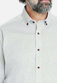 Camisa de botones en blanco, con un patrón geométrico en gris claro y negro. El cuello está estructurado con botones marrones para el cierre.