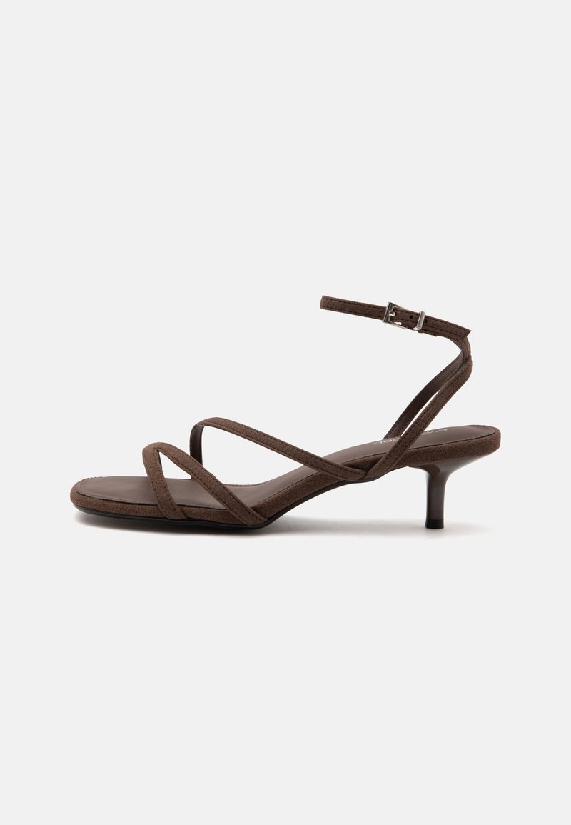 Sandale marron à talons hauts avec fines lanières croisées et bride de cheville avec boucle argentée sur fond blanc.