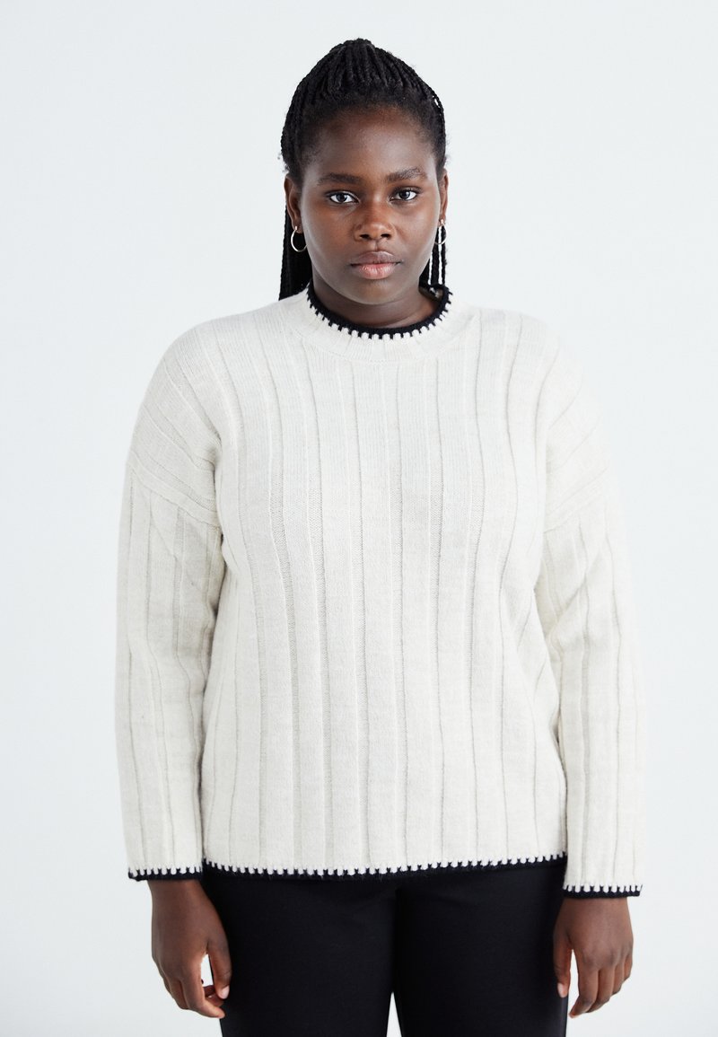 Cream-colored sweater with vertical ribbing, round neckline, and black pom-pom trim along the collar and hem, made from a soft material.