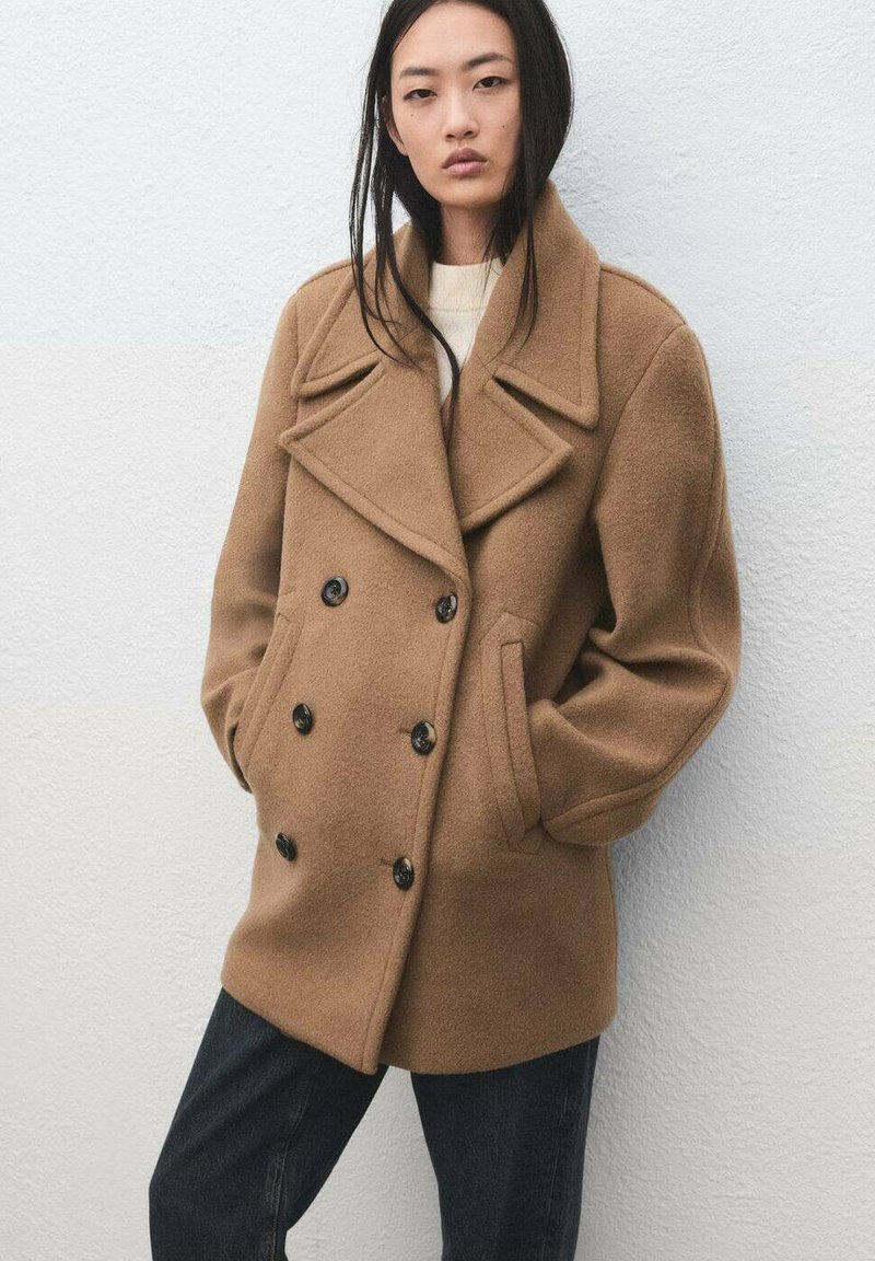 Cappotto doppiopetto in lana marrone, caratterizzato da grandi rever e bottoni neri. Il modello indossa jeans scuri su uno sfondo chiaro.