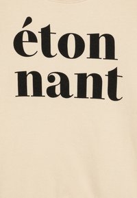 Beige džemperis ar lieliem melniem burtiem, kas lasa "étonnant." Audums izskatās gluds, ar klasisku apaļu kakla izgriezumu.