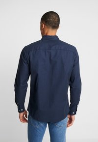 Námořnické modré košile s dlouhým rukávem a límcem na knoflíky, vyrobené z bavlny. Má manžety na jeden knoflík a mírně zakřivený lem.