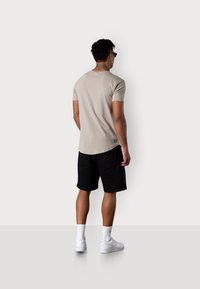 T-shirt beige à manches courtes associé à un short noir ; baskets blanches et chaussettes ; tatouage visible sur le cou ; détail du logo près de l'ourlet.