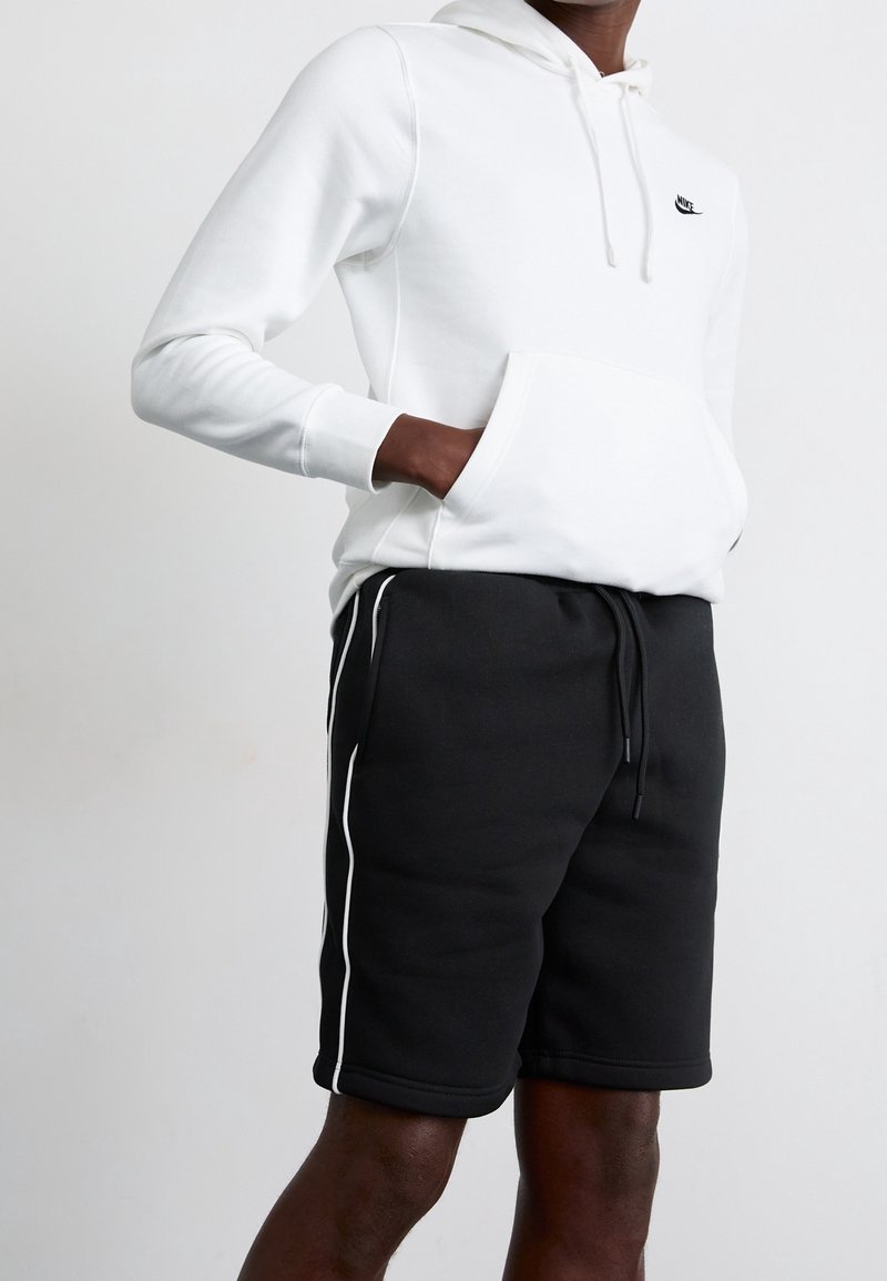 Personne portant un sweat à capuche blanc Nike avec poche avant et un short noir avec des bandes blanches sur les côtés sur un fond uni.