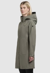 Cappotto lungo khaki con cappuccio, tasche laterali e zip frontale. Caratterizzato da una texture liscia e un design minimalista con un dettaglio del logo.