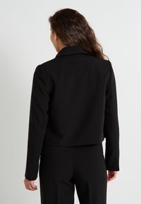 Mujer con cabello largo castaño, vistiendo una chaqueta negra corta y pantalones negros, de pie de espaldas contra un fondo blanco liso.