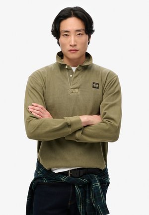 Olijfgroen polo met lange mouwen en een knoopkraag, geribbelde manchetten en een logo-embleem. Draagt een blauw en groen geruit shirt dat om de taille is geknoopt.