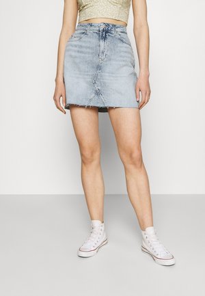 Kvinde iført lyseblå denim miniskørt med rå kant, blomsterprint crop top og hvide høje sneakers foran en ensfarvet baggrund.