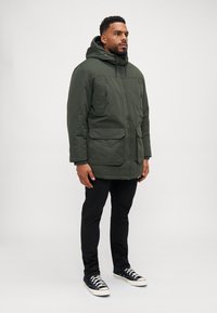 Chaqueta acolchada verde con capucha, dos bolsillos frontales y puños ajustables. Combinada con pantalones negros y zapatillas azul marino. Diseño sencillo y funcional.