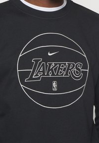 Sudadera negra con un diseño de baloncesto bordado en blanco, que muestra la palabra "LAKERS" y el logo de Nike. Textura de algodón suave.