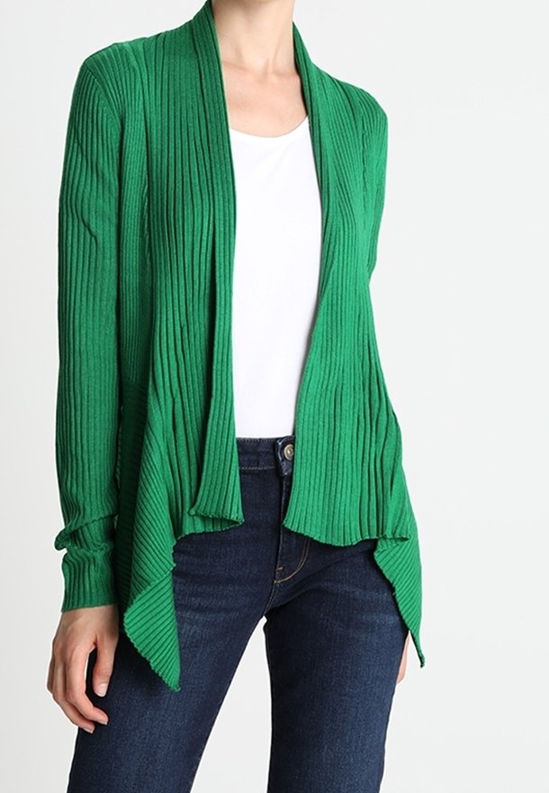 Personne portant un cardigan vert côtelé ouvert sur un haut blanc et un jean bleu foncé, debout devant un fond uni.