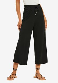 Črne hlače širokega kroja z vrvico v pasu, izdelane iz mehkega blaga, v kombinaciji s črnimi sandali z naramnicami na golih nogah.