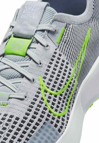 Zapatilla deportiva gris claro con parte superior de malla, acentos en verde y patrones texturizados. Presenta una marca visible y una suela blanca plana.
