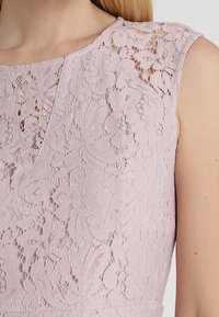 Robe en dentelle de couleur rose clair, avec un motif floral et un décolleté légèrement festonné. Conception sans manches avec un détail en dentelle texturée.