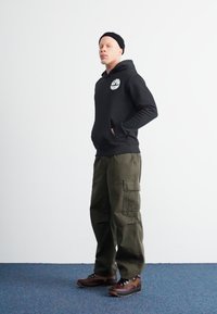 Hoodie preto com logotipo, calças cargo verdes-oliva e sapatos de caminhadas castanhos. Modelo está de pé contra um fundo plain com as mãos nos bolsos.