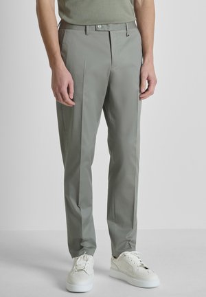 Hombre que lleva pantalones a medida gris claro y zapatillas blancas, de pie frente a un fondo liso y claro.