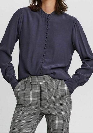 Femme portant un chemisier bleu marine foncé à manches longues boutonné, rentré dans un pantalon à carreaux noirs et blancs taille haute, les mains dans les poches.