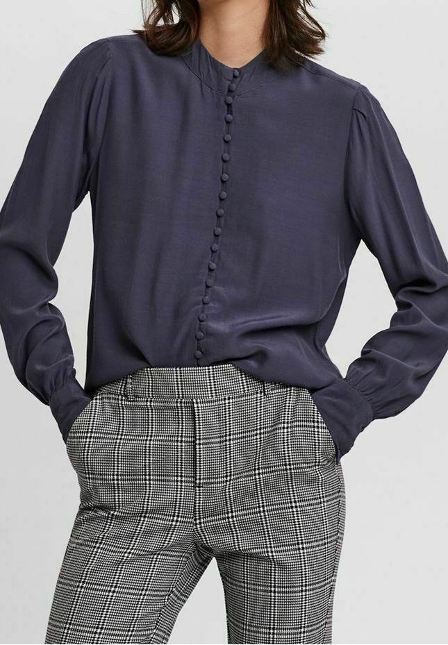 Femme portant un chemisier bleu marine foncé à manches longues boutonné, rentré dans un pantalon à carreaux noirs et blancs taille haute, les mains dans les poches.