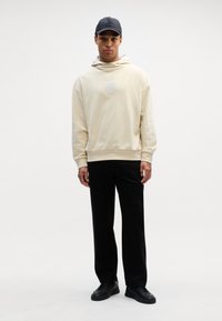 Beżowa bluza z kapturem z logo z przodu, zestawiona z czarnymi spodniami. Model nosi czapkę w kolorze czarnym oraz eleganckie czarne buty, stojąc na neutralnym tle.