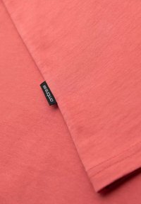 Bavlněné tričko v korálově růžové barvě s hladkou texturou. Má černou značkovou etiketu přišitou na lemu, která ukazuje jednoduchý design.