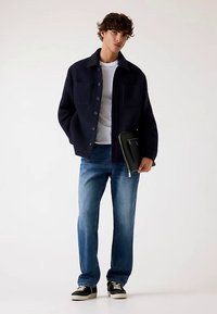 Giacca blu navy in misto lana con due tasche frontali, indossata sopra una t-shirt bianca, abbinata a jeans di denim blu e sneakers nere, mentre tiene in mano una custodia per tablet nera.