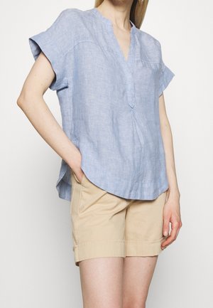 Mujer con blusa de lino azul claro de manga corta y escote con abertura, y pantalones cortos beige hasta la rodilla, de pie con una mano en el bolsillo.