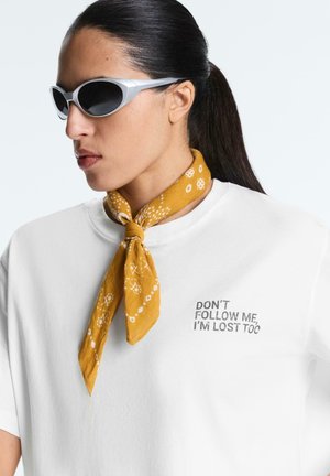 Žena s tmavými vlasy svázanými do culíku nosí bílé sluneční brýle, žlutý šátek se vzorem kolem krku a bílé tričko s nápisem „NEKOPÍRUJTE MĚ, TAKÉ JSEM ZTRACENÁ.“