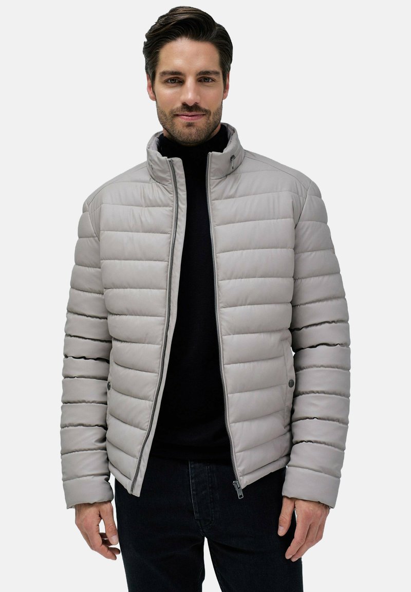 Chaqueta acolchada gris claro con un cuello alto, costuras horizontales, dos bolsillos con cremallera y un diseño ajustado. Usada sobre una camiseta negra.