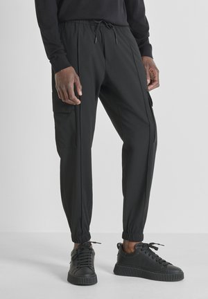 Man wearing black cargo jogger pants and black low-top sneakers, standing against a plain light background.