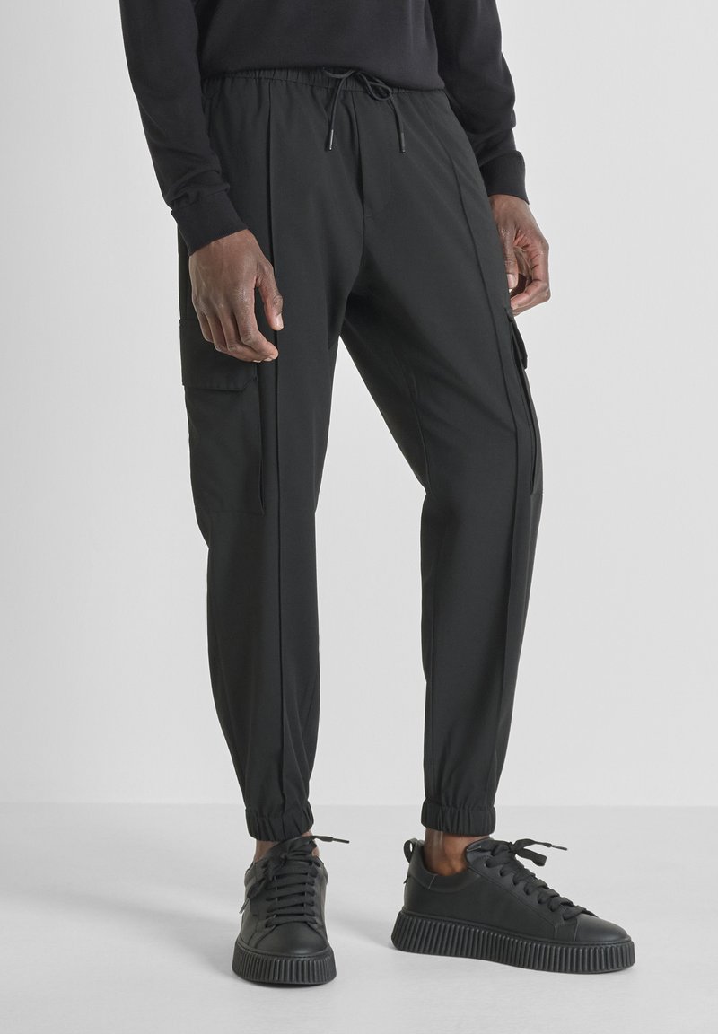 Man in zwarte cargo joggerbroeken en zwarte lage sneakers, staande tegen een effen lichte achtergrond.