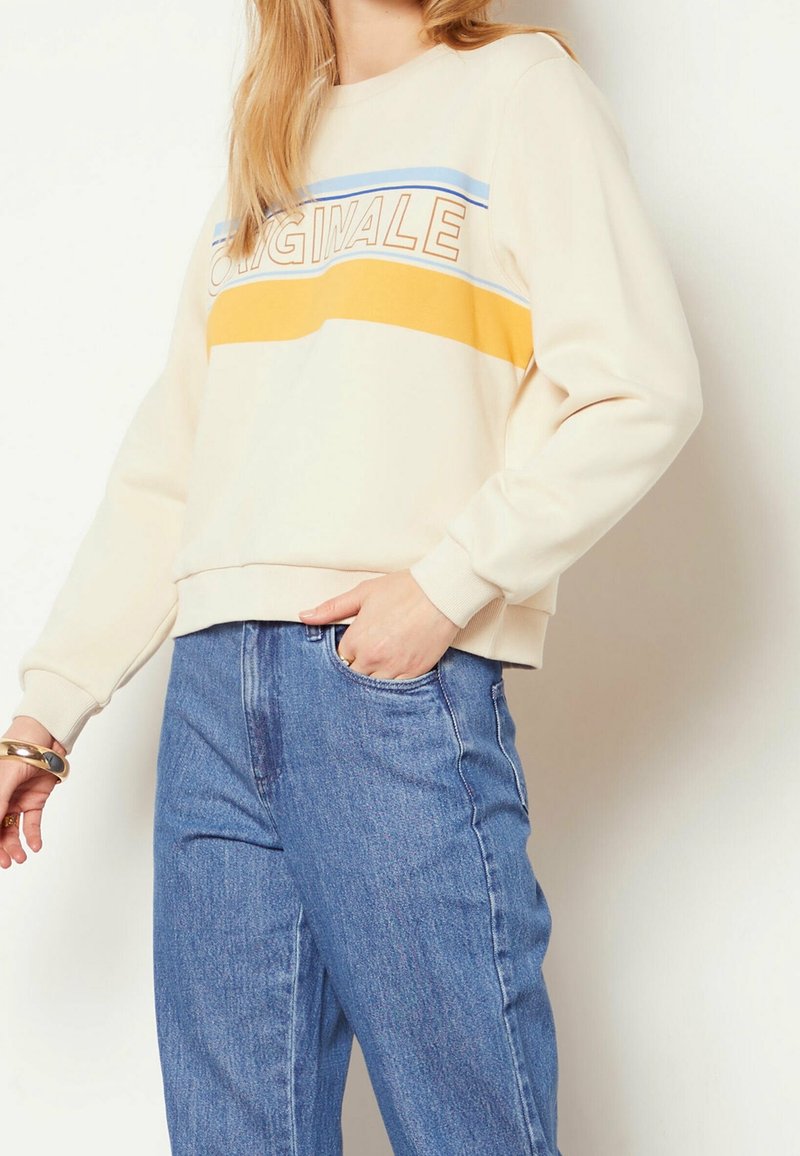 Personne portant un sweat crème à rayures bleues et jaunes avec le mot "ORIGINALE", associé à un jean bleu, main dans la poche.