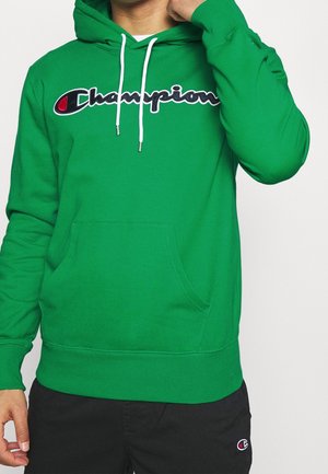 Mężczyzna w zielonej bluzie Champion z białymi sznurkami i czarnych spodniach z małym logo na udzie na jednolitym tle.