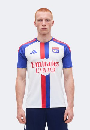 Mężczyzna ubrany w koszulkę piłkarską Olympique Lyonnais z białą bazą, niebiesko-czerwonymi pionowymi paskami oraz logo sponsora „Emirates Fly Better”.