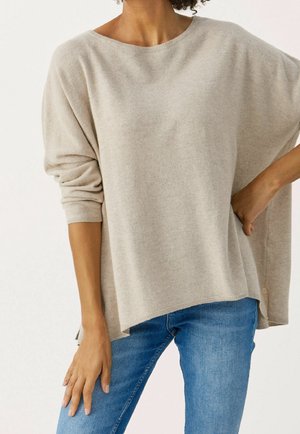 Persoon die een loszittende beige trui met lange mouwen draagt, gecombineerd met blauwe spijkerbroek, staand met één hand op de heup en de andere ontspannen langs het lichaam.