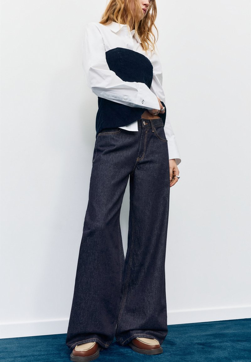 Oseba, oblečena v temne široke kavbojke, rjave čevlje, belo srajco z dolgimi rokavi in črno top brez naramnic, stoji ob beli steni na modrem tlaku.