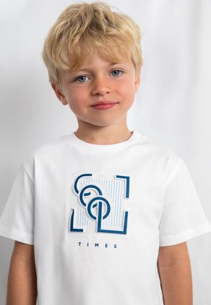 Ragazzo biondo con occhi azzurri che indossa una maglietta bianca con una grafica blu e la parola "TIMES" su uno sfondo uniforme.