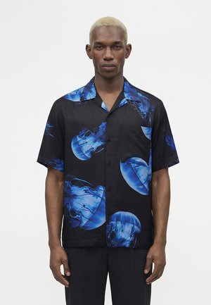 Mand med kort blondt hår iført en sort kortærmet skjorte med blå vandmønstre, står mod en almindelig hvid baggrund.