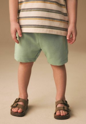 Barn står iført grønne shorts, stribet skjorte og brune sandaler med spænder på en neutral baggrund.