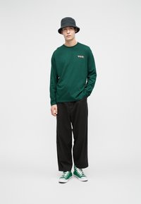 Młody mężczyzna w zielonej koszuli z długim rękawem, czarnych szerokich spodniach, zielonych trampkach i czarnym kapeluszu typu bucket, stojący na białym tle.