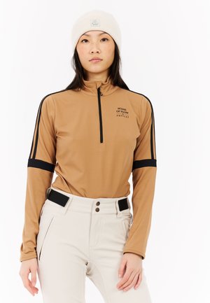 Light brown long-sleeve top with a quarter-zip, featuring black stripes on the arms and a logo. Paired with white pants with two buttons.
