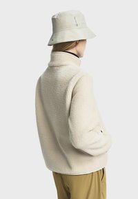 Chaqueta de borreguillo beige con cuello alto, superficie texturada y mangas largas. Combinada con un sombrero de cubo de color claro.