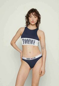 Kobieta w niebiesko-białym dwuczęściowym stroju kąpielowym Tommy Jeans, stojąca z jedną ręką na biodrze na tle jasnego, jednolitego tła.