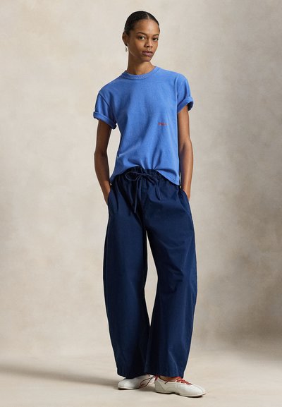 Μπλε βαμβακερό t-shirt με γυρισμένα μανίκια και κόκκινο λογότυπο, συνδυασμένο με φαρδιά σκούρα μπλε παντελόνια. Το μοντέλο στέκεται με τα χέρια στις τσέπες, φοράει λευκά παπούτσια.