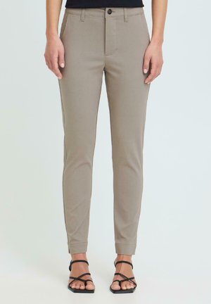 Pantalon beige slim avec bouton et fermeture éclair à l'avant, porté avec des sandales noires à talons et à lanières, présenté sur une personne debout sur un fond uni.
