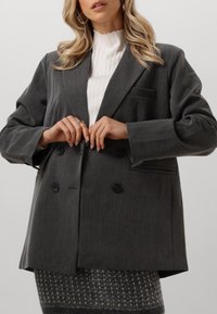 Harmaa kaksirivinen bleiseri, jossa on teksturoitu kangas, suuret mustat napit ja yksi rintatasku. Yhdistetty valkoisen ribatun paidan kanssa.